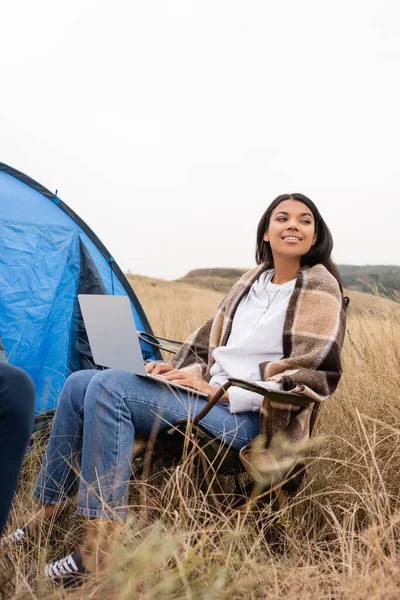 Sorridente donna africana americana avvolta in una coperta utilizzando il computer portatile vicino tenda sul prato — Foto stock