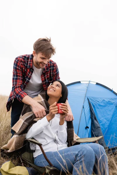 Lächelnder Mann legt beim Zelten auf dem Rasen Decke über afrikanisch-amerikanische Freundin mit Tasse in Zeltnähe — Stockfoto