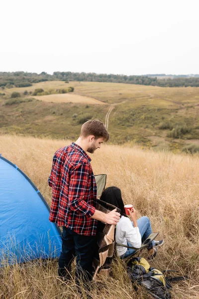 Man holding blanket near african american girlfriend with cup and tent on grassy field — Stock Photo