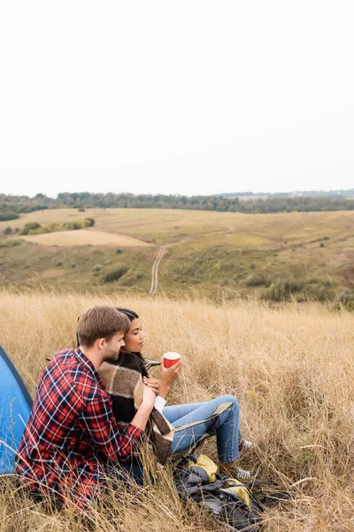 Hombre abrazando afroamericano novia con taza durante camping en campo herboso - foto de stock