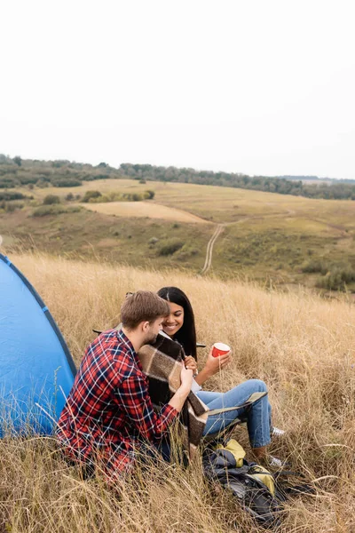 Coppia multietnica con coperta e coppa seduta vicino allo zaino e alla tenda sul campo — Foto stock