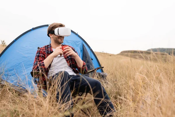 Smiling man in vr headset holding cup near tent on grassy meadow — Stock Photo