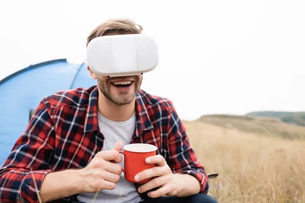 Hombre sonriente con auriculares vr sosteniendo la taza cerca de la tienda sobre fondo borroso en el prado - foto de stock