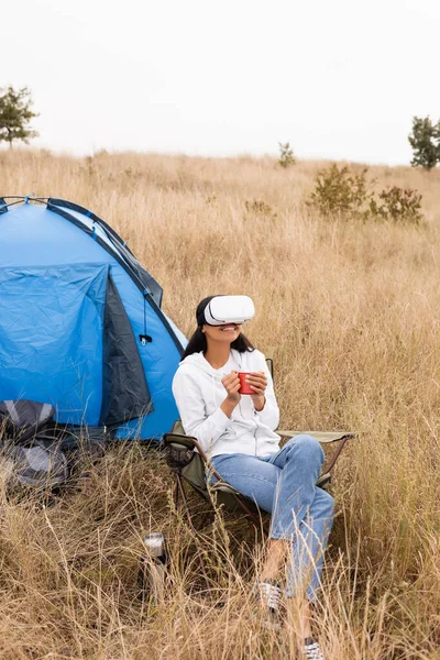 Sourire femme afro-américaine dans vr casque tenant tasse près de la tente sur pelouse herbeuse — Photo de stock