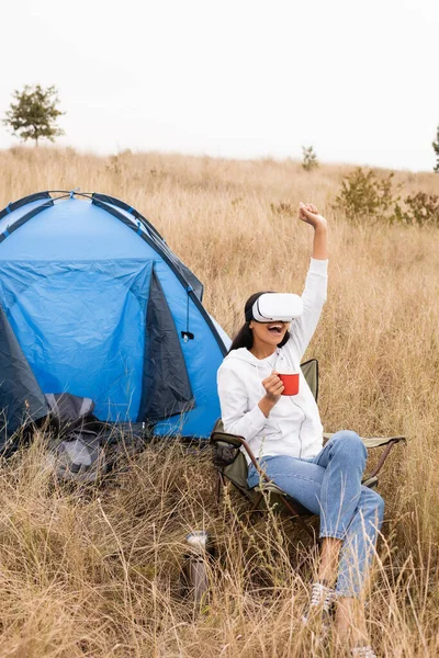 Allegro donna africana americana in auricolare vr tenendo tazza durante il campeggio vicino tenda sul prato — Foto stock