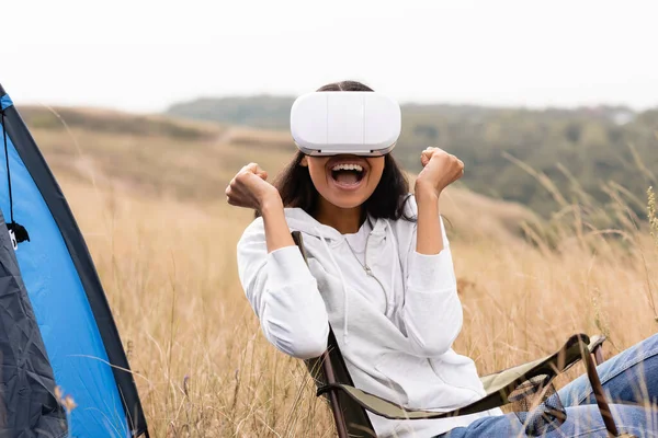 Femme afro-américaine joyeuse utilisant un casque de réalité virtuelle sur une chaise près de la tente sur le terrain — Photo de stock