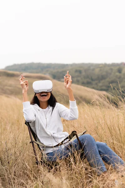 Alegre mujer afroamericana con dedos cruzados usando auriculares vr en silla en el campo - foto de stock