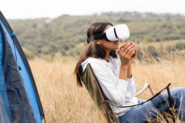 Sonriente mujer afroamericana en vr auriculares sosteniendo taza mientras está sentado en la silla cerca de la tienda en el prado - foto de stock