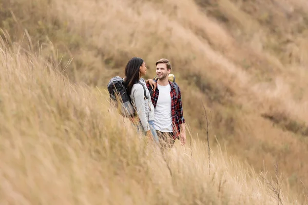 Couple interracial souriant avec des sacs à dos se regardant sur une colline gazeuse — Photo de stock