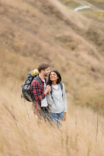 Усміхнена багатоетнічна пара з рюкзаками тримає руки на трав'янистому пагорбі — стокове фото