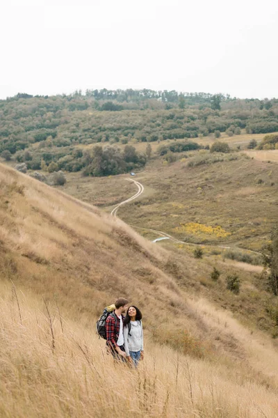 Усміхнена міжрасова пара з рюкзаками, що стоять на пагорбі з пейзажем на фоні — стокове фото