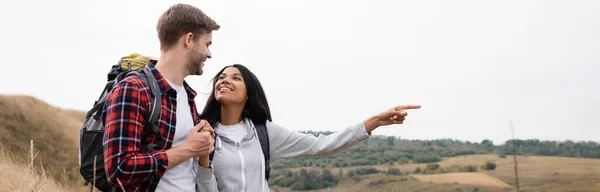 Sonriente mujer afroamericana señalando con el dedo y sosteniendo la mano del novio durante el viaje, pancarta - foto de stock