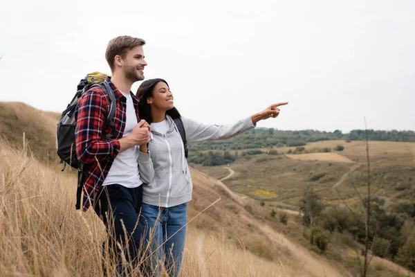 Smiling african american woman pointing with finger near boyfriend during trip with backpacks — Stock Photo