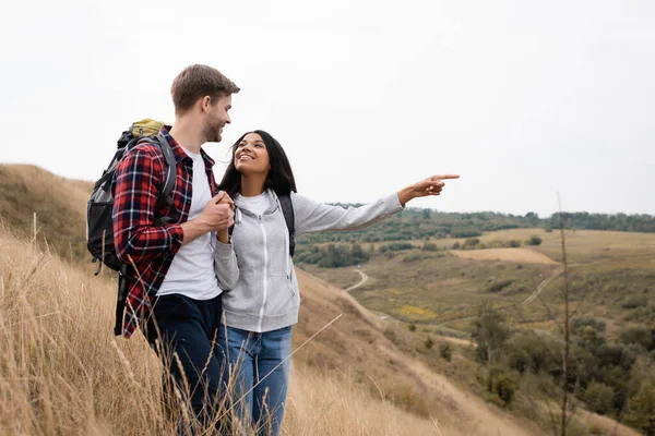 African american woman with backpack pointing with finger at landscape near boyfriend with backpack — Stock Photo