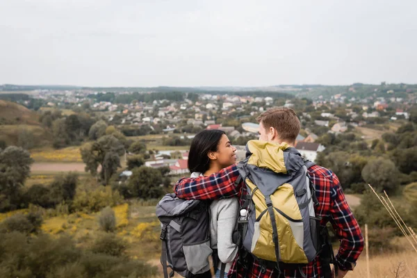 Back view of man embracing african american girlfriend with backpack during trip outdoors — Stock Photo