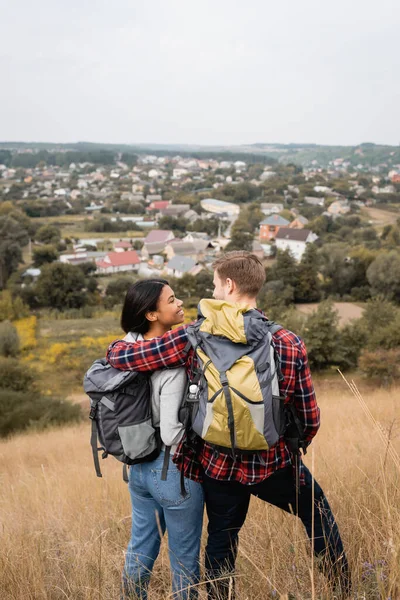Rückansicht von Tourist mit Rucksack umarmt lächelnde afrikanisch-amerikanische Freundin auf grasbewachsenem Hügel — Stockfoto