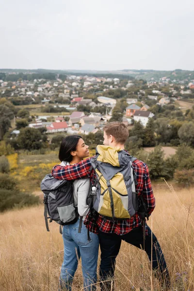 Vista trasera de turista con mochila abrazando sonriente novia afroamericana en colina cubierta de hierba - foto de stock