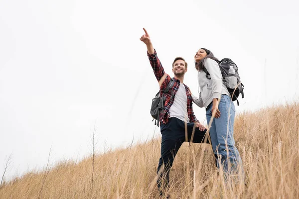 Low angle view of smiling man with backpack pointing with finger away near african american woman while standing on grassy hill with sky at background — Stock Photo