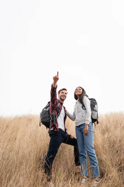 Vista de ángulo bajo del turista afroamericano sonriente parado cerca de novio señalando con el dedo en la colina cubierta de hierba — Stock Photo