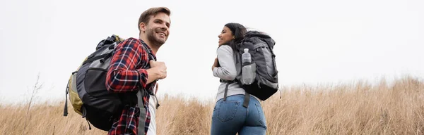 Smiling multiethnic hikers with backpacks walking on hill with sky at background, banner — Stock Photo