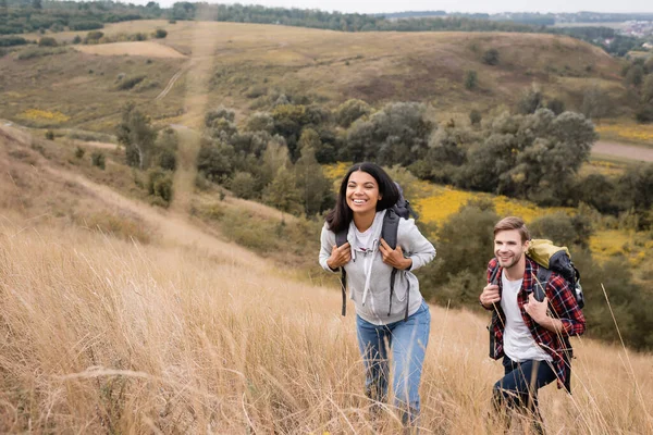 Interrassische Paar mit Rucksäcken zu Fuß auf Hügel mit Gras während der Reise — Stockfoto