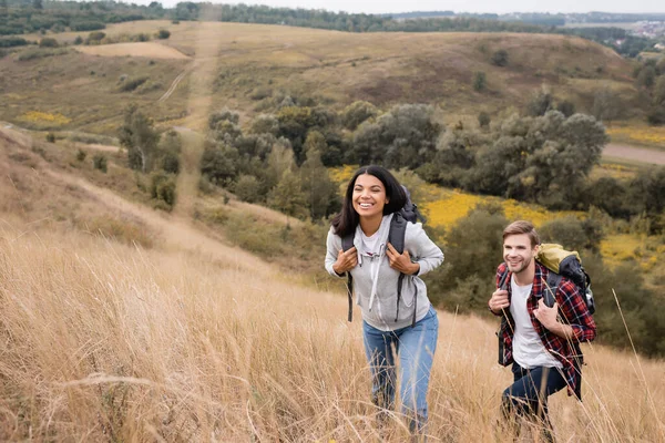 Interracial couple with backpacks walking on hill with grass during trip — Stock Photo
