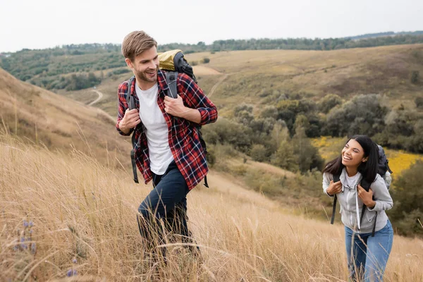 Усміхнений чоловік з рюкзаком, що йде біля афроамериканської дівчини на пагорбі з травою — стокове фото