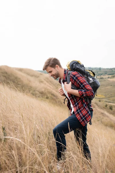 Sonriente hombre caminando con mochila en la colina con hierba - foto de stock