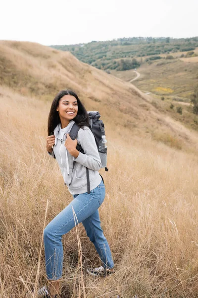 Positivo escursionista afro-americano che tiene lo zaino mentre cammina sulla collina con erba — Foto stock