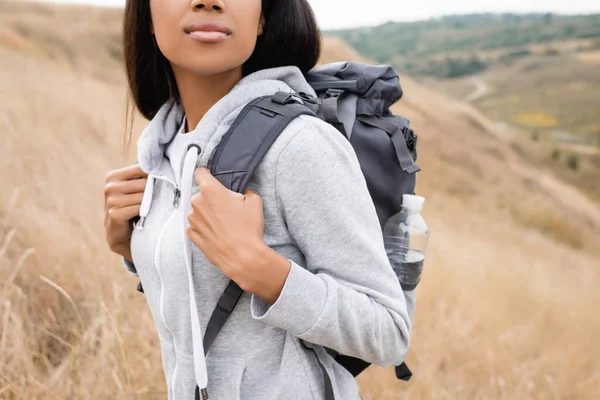 Vue recadrée d'une femme afro-américaine avec un sac à dos debout sur une colline avec un paysage flou en arrière-plan — Photo de stock