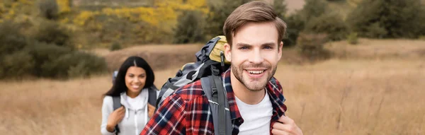 Smiling traveler with backpack looking away near african american girlfriend on blurred background during trip, banner — Stock Photo
