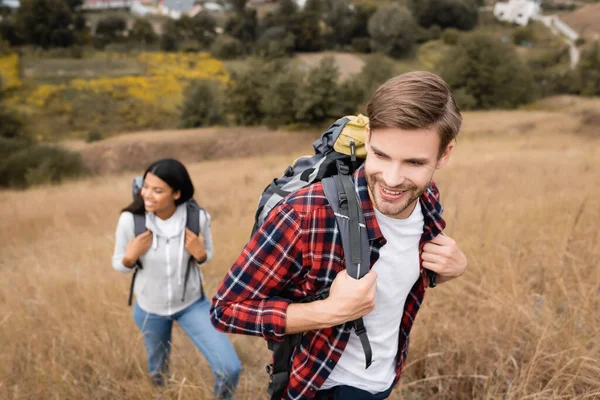 Sorridente turista toccante zaino vicino afro americano donna su sfondo sfocato sulla collina — Foto stock