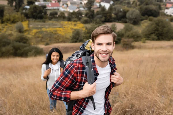 Cheerful traveler looking at camera near african american girlfriend on blurred background during trip — Stock Photo