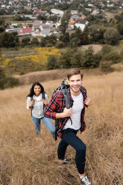 Молодий турист з рюкзаком посміхається на камеру біля афроамериканки на розмитому фоні на відкритому повітрі — стокове фото