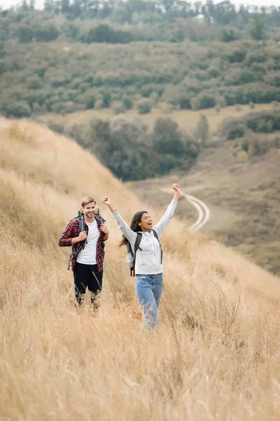 Cheerful african american traveler with backpack showing yeah gesture near boyfriend on field — Stock Photo
