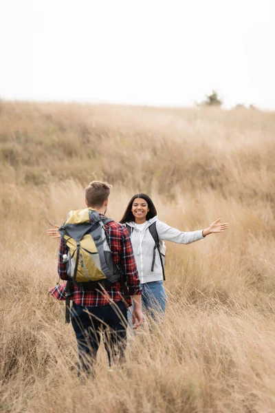 Smiling african american woman smiling at boyfriend with backpack on blurred foreground on grassy hill — Stock Photo