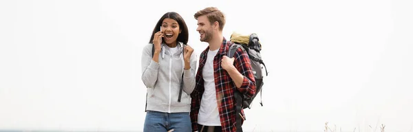 Fröhliche afrikanisch-amerikanische Frau spricht auf Smartphone neben Freund mit Rucksack isoliert auf grau, Banner — Stockfoto