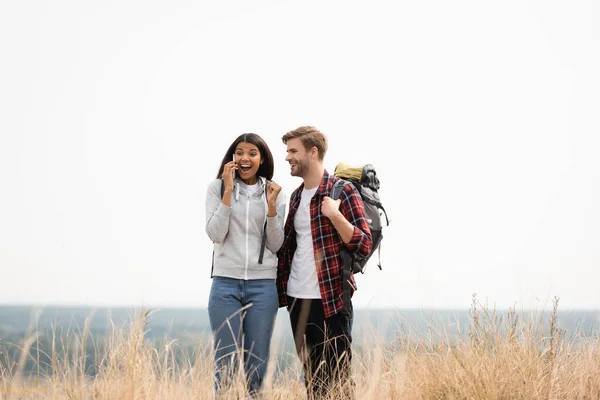 Alegre mujer afroamericana hablando en teléfono inteligente cerca de novio con mochila durante el viaje - foto de stock