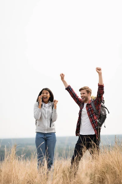 Lächelnder Reisender zeigt Ja-Geste nahe afrikanisch-amerikanischer Freundin, die im Freien auf Smartphone spricht — Stockfoto