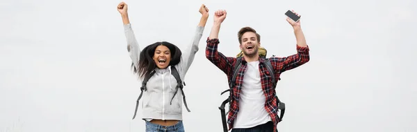 Cheerful multiethnic couple with backpacks and smartphone isolated on grey, banner — Stock Photo