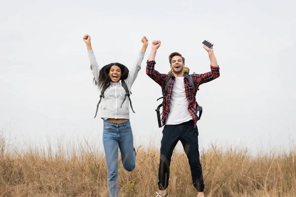 Positive multiethnische Reisende mit Rucksack und Smartphone, die auf dem Rasen in die Kamera schauen — Stockfoto