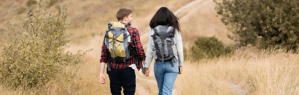 Vista posterior de la joven pareja de viajeros con mochilas caminando en el camino al aire libre, pancarta - foto de stock
