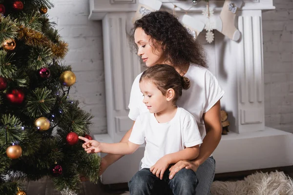 Madre e hija tocando la bola de Navidad en el pino festivo cerca de la chimenea - foto de stock