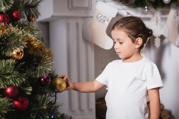 Fille debout près de la cheminée et touchant boule de Noël sur le pin festif à la maison — Photo de stock