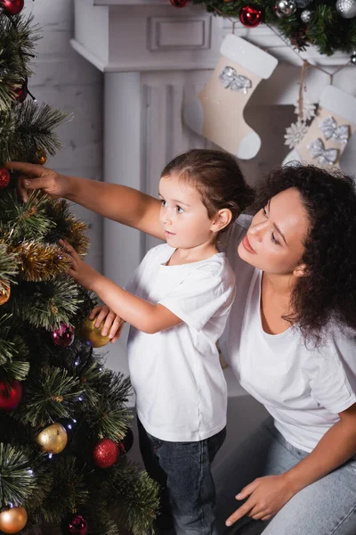 Madre e hija decorando pino de Navidad cerca de la chimenea en casa - foto de stock