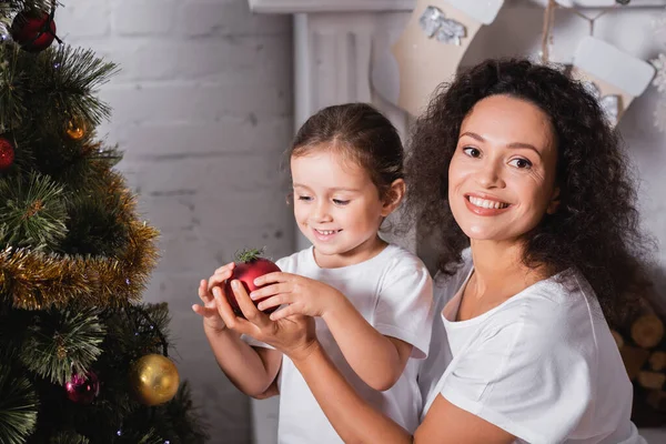 Mother and daughter with christmas ball near festive pine at home — Stock Photo