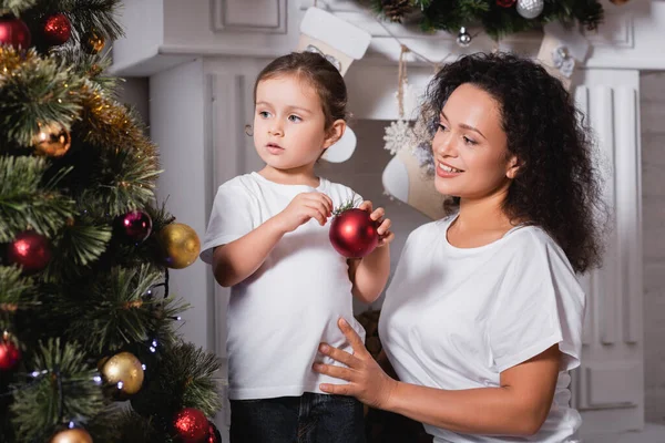 Mother and daughter with christmas ball standing near festive pine and fireplace — Stock Photo