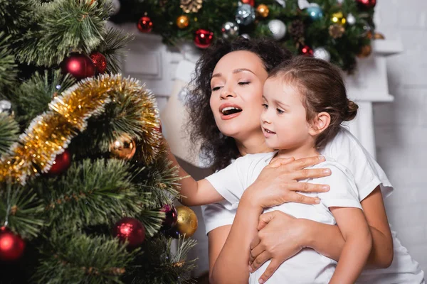 Mutter umarmt Tochter bei dekorierter Kiefer zu Hause — Stockfoto