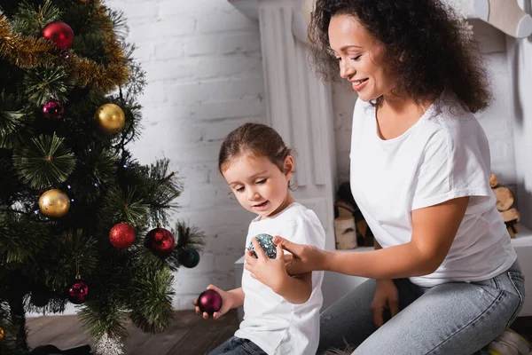 Hija y madre con bolas de Navidad cerca de pino festivo en casa - foto de stock