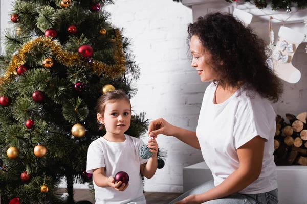 Madre e hija con bolas de Navidad cerca de pino decorado y chimenea - foto de stock